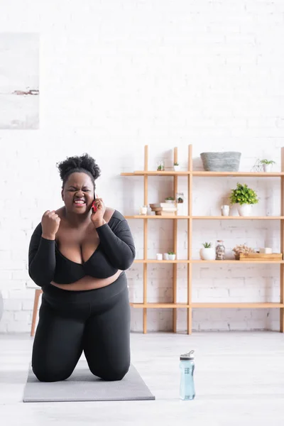 Erstaunte afrikanisch-amerikanische Plus-Size-Frau in Sportbekleidung, die auf der Fitnessmatte sitzt und mit dem Smartphone spricht — Stockfoto