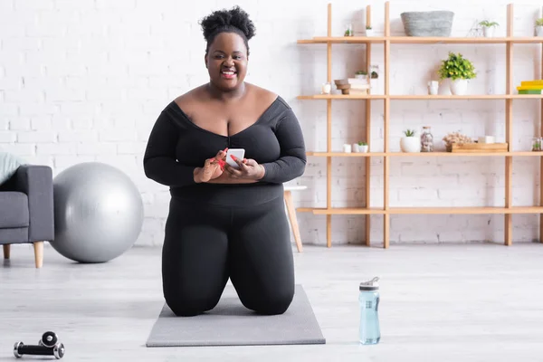 Happy african american plus size woman in sportswear using smartphone and standing on knees on fitness mat — Stock Photo