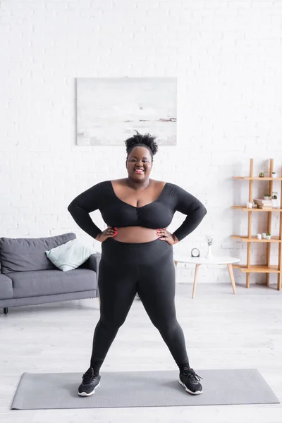 Pleine longueur de femme afro-américaine heureuse en surpoids en vêtements de sport debout avec les mains sur les hanches à la maison — Photo de stock