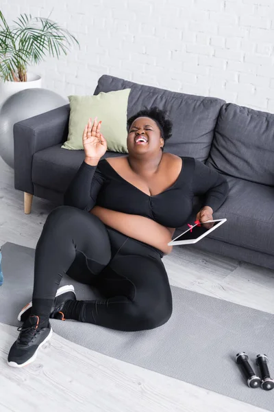 Fröhliche afrikanisch-amerikanische Plus-Size-Frau in Sportbekleidung mit digitalem Tablet und lachendem Sofa im Wohnzimmer — Stockfoto