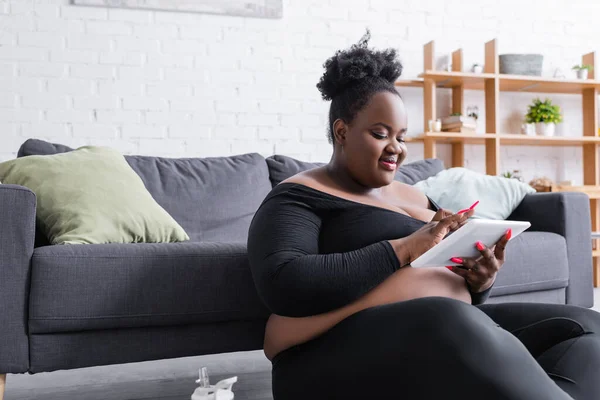 Pleased african american plus size woman in sportswear using digital tablet in living room — Stock Photo
