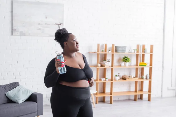 Fröhliche afrikanisch-amerikanische übergewichtige Frau in Sportbekleidung mit Sportflasche zu Hause — Stockfoto