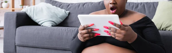 Vue recadrée de femme afro-américaine choquée plus la taille dans les vêtements de sport en utilisant une tablette numérique dans le salon, bannière — Photo de stock