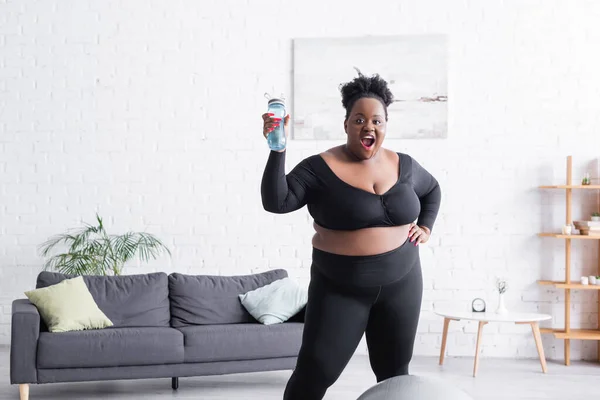 Espantado afro-americano plus size mulher em sportswear segurando garrafa de esportes em casa — Fotografia de Stock