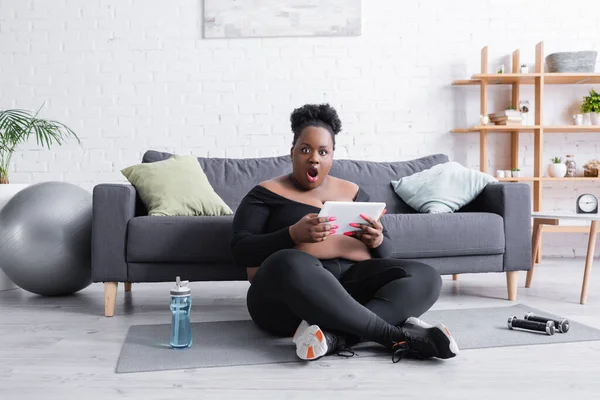 Afro-américaine choquée plus taille femme tenant tablette numérique dans le salon — Photo de stock