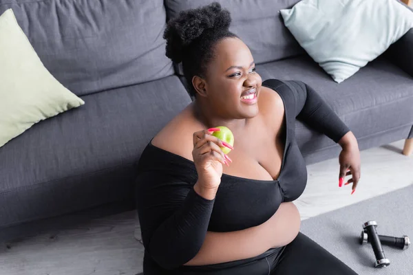 Cheerful african american plus size woman in sportswear holding green apple — Stock Photo