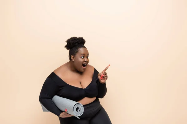 Étonné afro-américain plus taille femme en tenue de sport tenant tapis de fitness et pointant avec le doigt isolé sur beige — Photo de stock