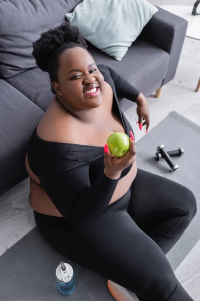 Vue grand angle de heureux afro-américain plus taille femme en tenue de sport tenant pomme verte — Photo de stock