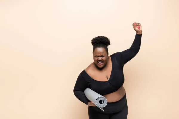 Étonné afro-américain plus taille femme en tenue de sport tenant tapis de fitness isolé sur beige — Photo de stock