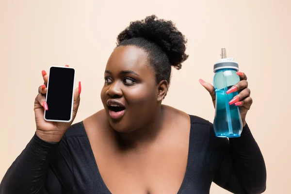 Étonné afro-américain plus taille femme tenant bouteille de sport et smartphone avec écran blanc isolé sur beige — Photo de stock