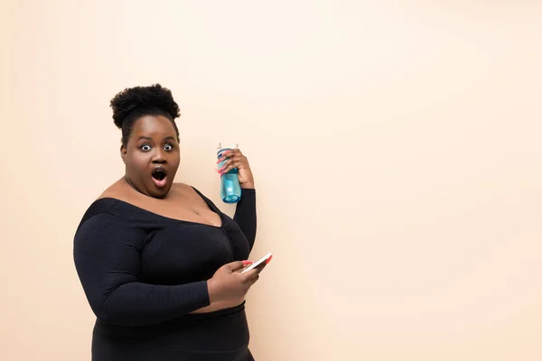 Choqué afro-américain plus taille femme en tenue de sport tenant bouteille de sport et smartphone isolé sur beige — Photo de stock