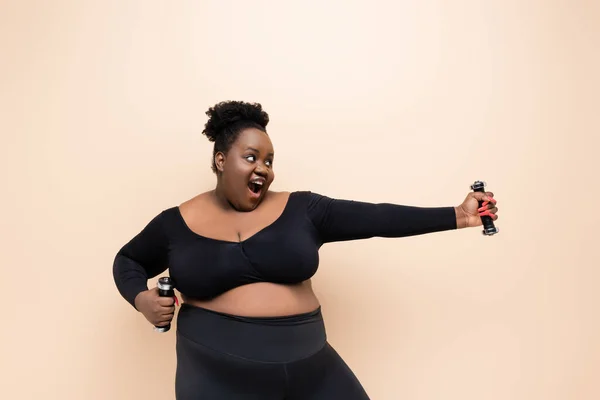 Aufgeregte afrikanisch-amerikanische Plus-Size-Frau in Sportbekleidung mit Hanteln isoliert auf beige — Stockfoto