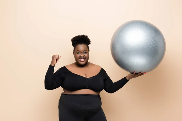Fröhliche afrikanisch-amerikanische Plus-Size-Frau in Sportbekleidung hält Fitnessball isoliert auf beige — Stockfoto