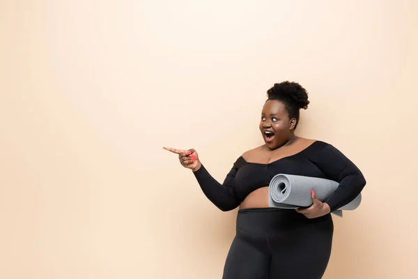 Erstaunt afrikanisch-amerikanische übergewichtige Frau in Sportbekleidung hält Fitnessmatte und zeigt mit dem Finger isoliert auf beige — Stockfoto