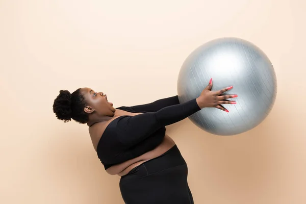 Überraschte afrikanisch-amerikanische Plus-Size-Frau in Sportbekleidung hält Fitnessball isoliert auf beige — Stockfoto