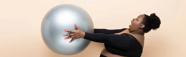 Überraschte afrikanisch-amerikanische Plus-Size-Frau in Sportbekleidung mit Fitnessball isoliert auf beige, Banner — Stockfoto