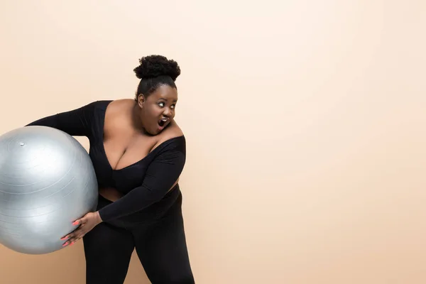 Schockierte afrikanisch-amerikanische Plus-Size-Frau in Sportbekleidung hält Fitnessball isoliert auf beige — Stockfoto