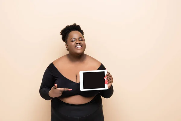 Heureux afro-américain plus taille femme pointant avec le doigt à la tablette numérique avec écran blanc isolé sur beige — Photo de stock