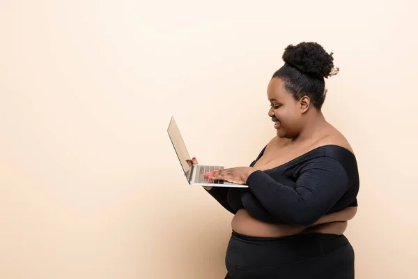 Side view of happy african american plus size woman holding laptop isolated on beige — Stock Photo