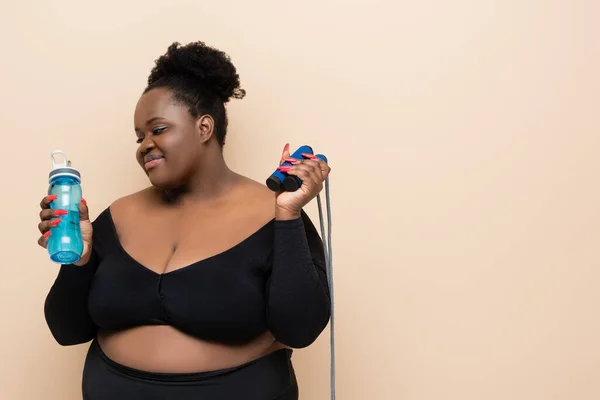 Glückliche afrikanisch-amerikanische Plus-Size-Frau in Sportbekleidung mit Sportflasche und Springseil isoliert auf beige — Stockfoto