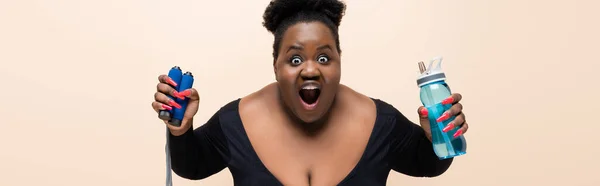 Schockierte afrikanisch-amerikanische Plus-Size-Frau mit Sportflasche und Springseil isoliert auf beige, Banner — Stockfoto