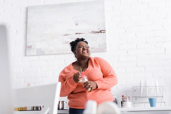 Gai afro-américain plus taille femme tenant tasse en plastique avec smoothie savoureux dans la cuisine — Photo de stock