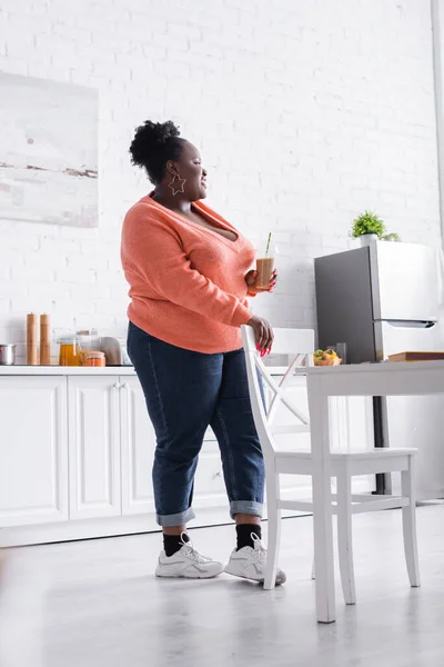 Pleased african american plus size woman holding plastic cup with tasty smoothie in kitchen — Stock Photo