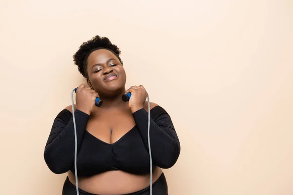 Pleased african american plus size woman in sportswear holding skipping rope isolated on beige — Stock Photo