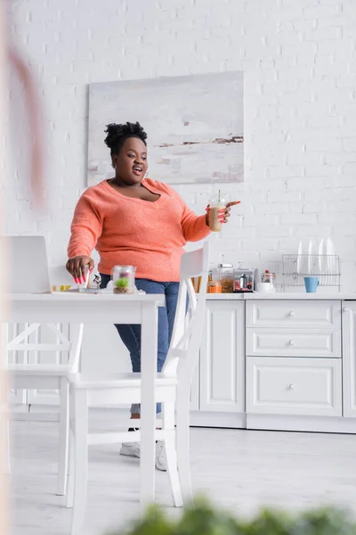 Heureux afro-américain plus taille femme tenant tasse en plastique avec smoothie dans la cuisine avec avant-plan flou — Photo de stock