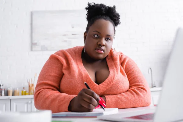 Lockige afrikanisch-amerikanische Plus-Size-Frau mit Laptop und Stift in der Nähe Notizbuch — Stockfoto
