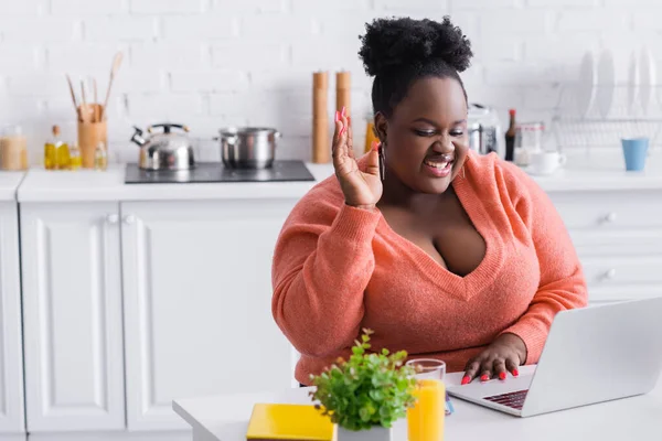 Feliz afroamericano más freelancer de tamaño con la mano levantada usando el ordenador portátil en casa - foto de stock