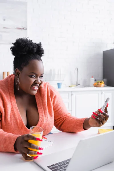 Heureux afro-américain plus taille femme tenant un verre de jus d'orange et regardant smartphone — Photo de stock