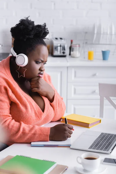 Afro-américaine plus femme taille écoute podcast dans les écouteurs et en utilisant un ordinateur portable — Photo de stock