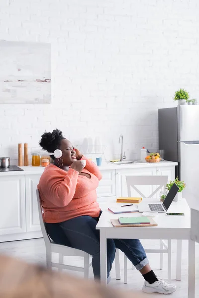 Happy african american plus size woman in headphones holding cup in kitchen — Stock Photo