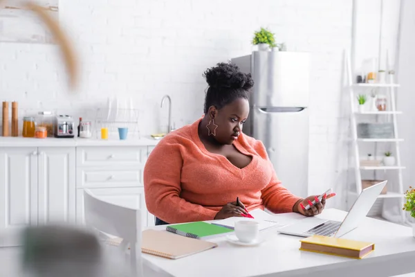 Afro-américaine plus taille femme tenant smartphone près d'un ordinateur portable sur la table — Photo de stock