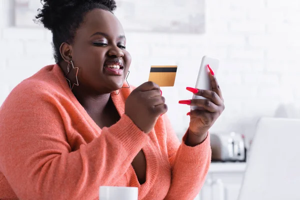 Souriant afro-américain plus taille femme tenant carte de crédit et téléphone mobile — Photo de stock