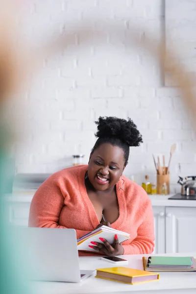 Glückliche afrikanisch-amerikanische Plus-Size-Frau schaut auf Laptop und hält Notizbuch — Stockfoto