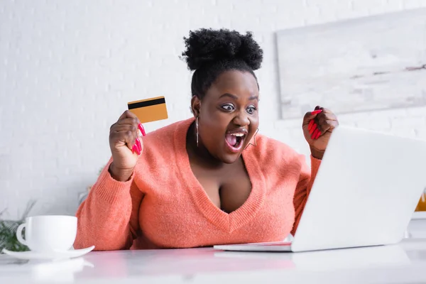 Étonné afro-américain plus la taille femme tenant la carte de crédit et regardant ordinateur portable — Photo de stock