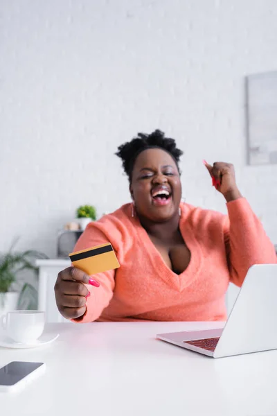 Feliz africano americano más tamaño mujer celebración de la tarjeta de crédito cerca de la computadora portátil - foto de stock