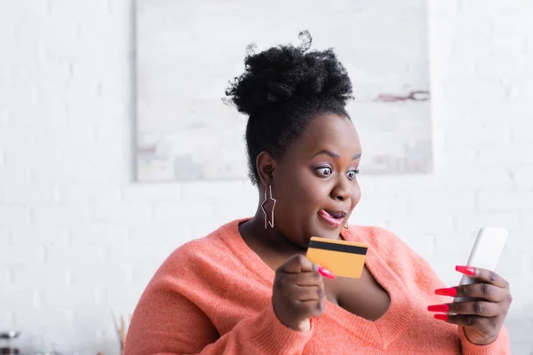 Afro-américaine plus taille femme qui sort la langue tout en tenant la carte de crédit et smartphone — Photo de stock