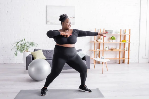 Volle Länge der zufriedenen afrikanisch-amerikanischen plus size Frau in Sportbekleidung, die im Wohnzimmer trainiert — Stockfoto