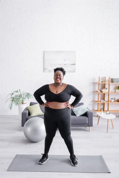 Volle Länge der afrikanisch-amerikanischen Plus-Size-Frau in Sportbekleidung stehend mit den Händen auf den Hüften auf der Fitnessmatte — Stockfoto