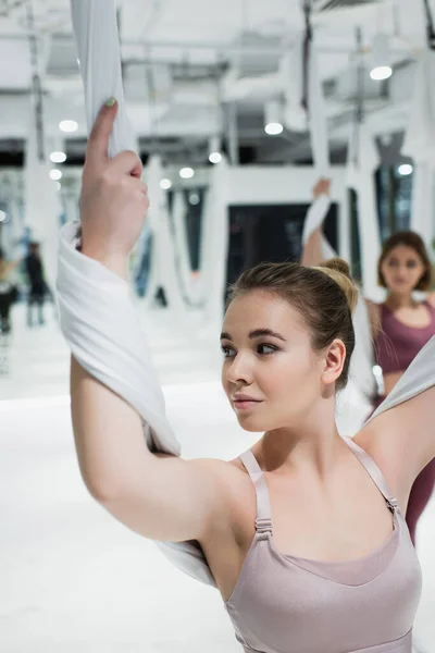 Pretty woman looking away while stretching arms with fly yoga strap, blurred background — Stock Photo