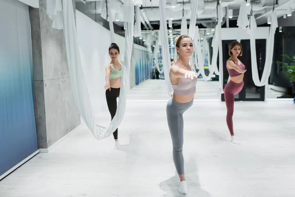 Sportliche Frauen üben Kriegerpose in der Nähe von Fliege Yoga-Hängematten in der Turnhalle — Stockfoto