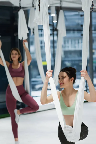 Lächelnde Frau wärmt sich mit Antenne Yoga-Hängematte auf verschwommenem Hintergrund auf — Stockfoto