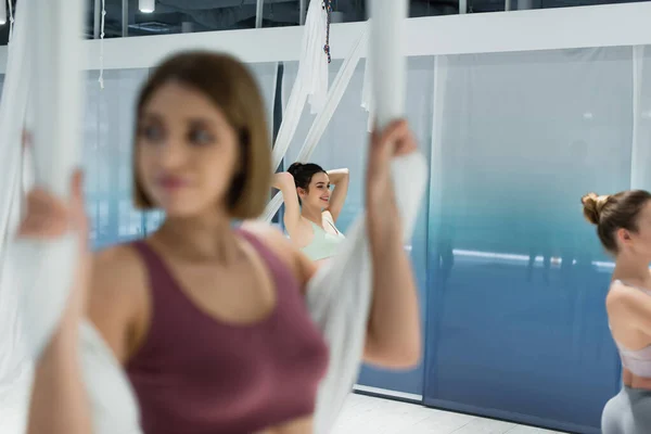 Вибірковий фокус усміхненої жінки, що розігрівається повітряним гамаком йоги на розмитому передньому плані — стокове фото