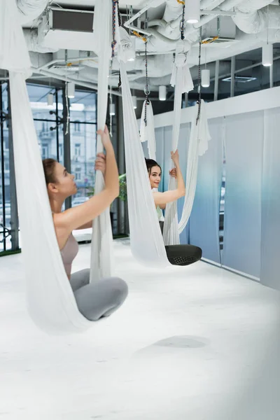 Young sportswomen sitting in lotus pose in aerial yoga hammocks, blurred foreground — Stock Photo