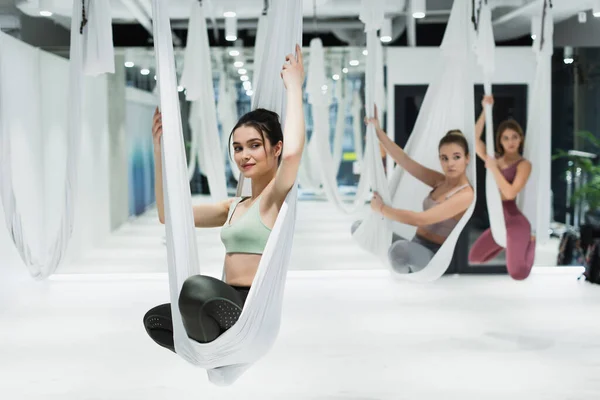 Mujer feliz que mira lejos mientras que practica la pose del loto en la hamaca del yoga de la mosca en fondo borroso - foto de stock