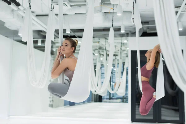 Mujer joven mirando hacia otro lado mientras que se sienta en la pose del loto en la hamaca aérea del yoga - foto de stock