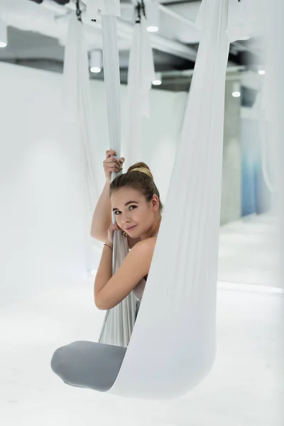 Mujer joven mirando la cámara mientras que está sentada en la hamaca aérea del yoga - foto de stock